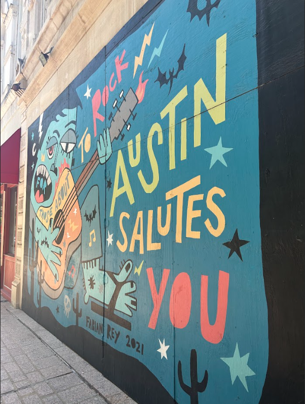 This image shows a blue wall mural found in Austin, TX featuring a cartoon character with a guitar and the words "For Those About to Rock, Austin Salutes You"